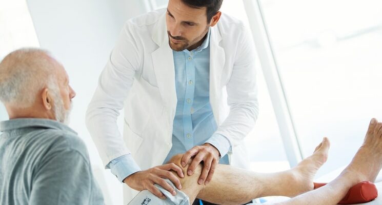 Closeup over the shoulder view of an early 60's senior gentleman having some knee pain. He's at doctor's office having medical examination by a male doctor. The doctor is touching the sensitive area and trying to determine the cause of pain.