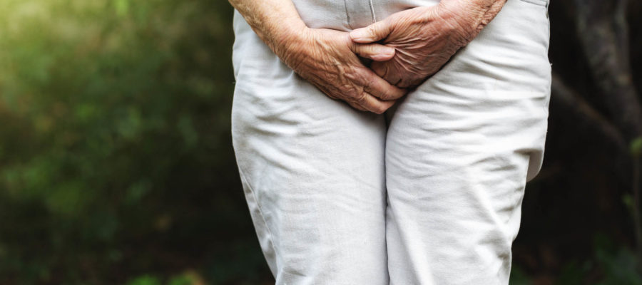 The wrinkled hands of an old woman press on her groin area through her trousers in an effort to prevent involuntary urination.